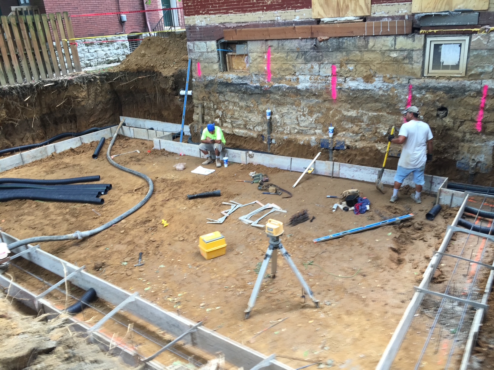 Crew laying out footings and drainage at an excavated foundation site beside an existing building